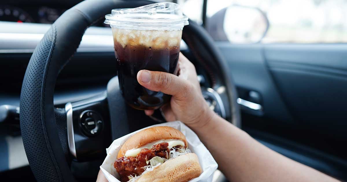 Photo showing a person's hands holding a sandwich and a drink while they are behind the wheel of a car.